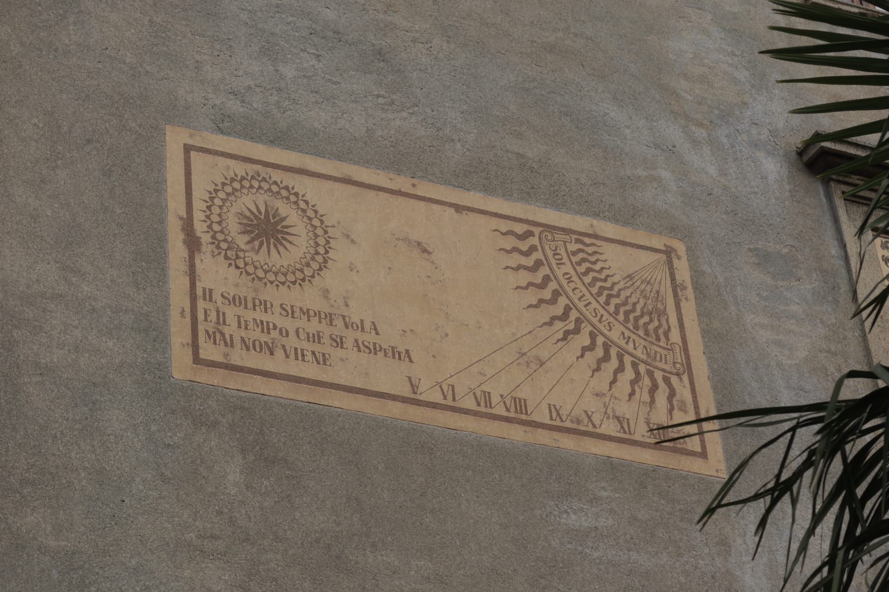 A large painted vertical sundial on the stucco wall of an Italian building, with ornate hour lines and decorative border.