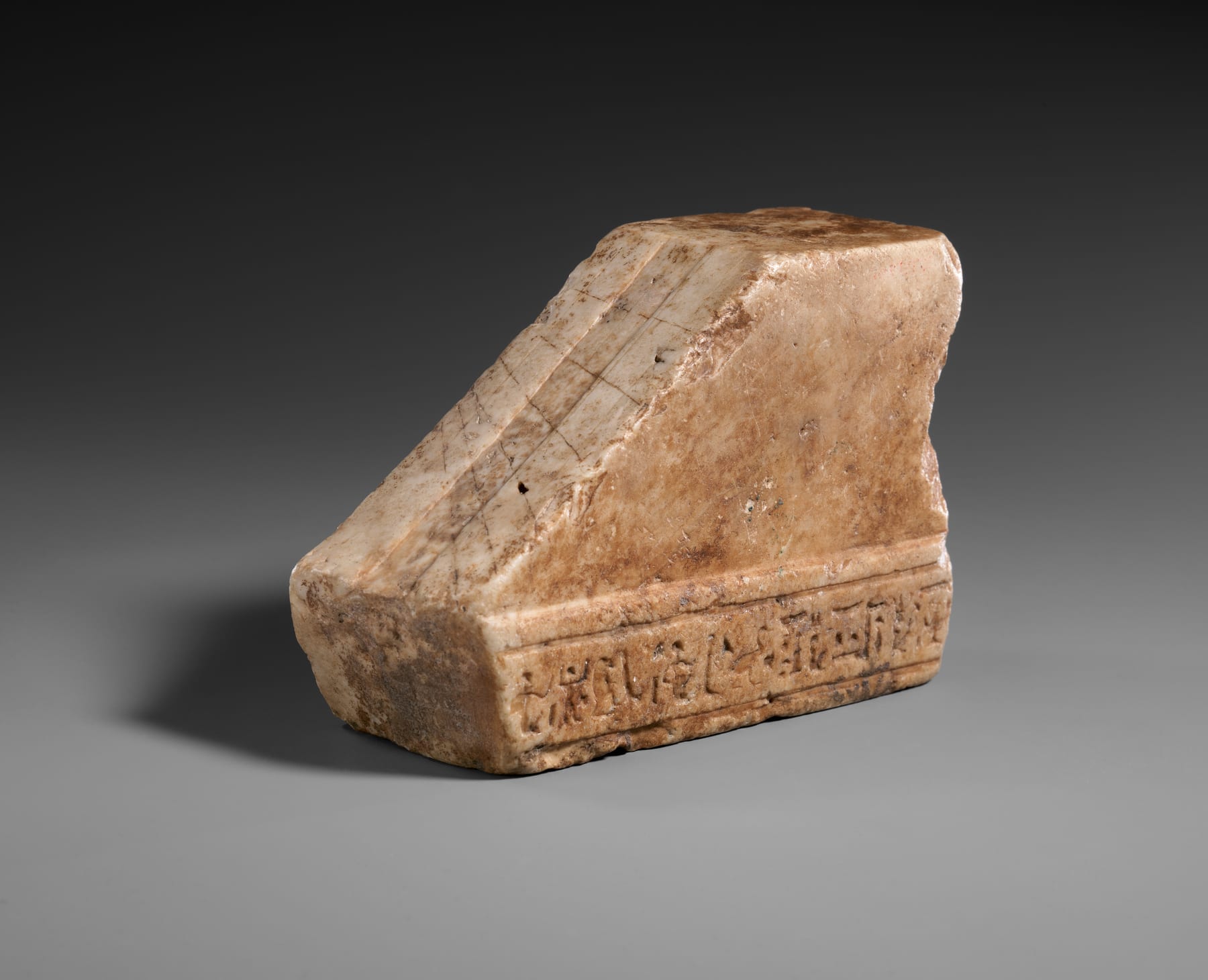 A small limestone ancient Egyptian shadow clock, hand-sized, with engraved tick marks along the horizontal arm and a central raised element that would cast a shadow across the scale.