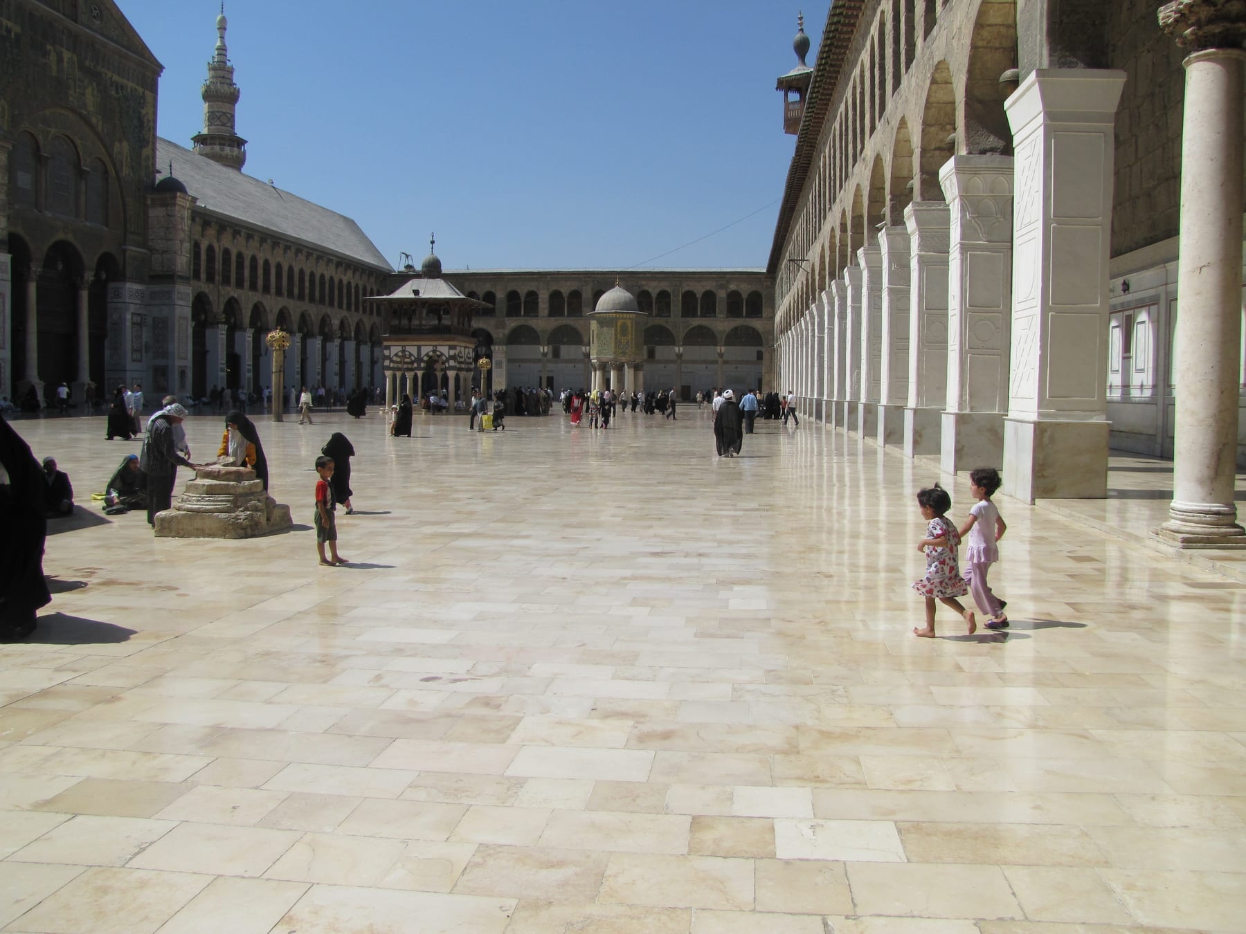 The large stone courtyard of the Umayyad Mosque in Damascus, flanked by tall arcades and with the mosque's minarets visible against a blue sky.